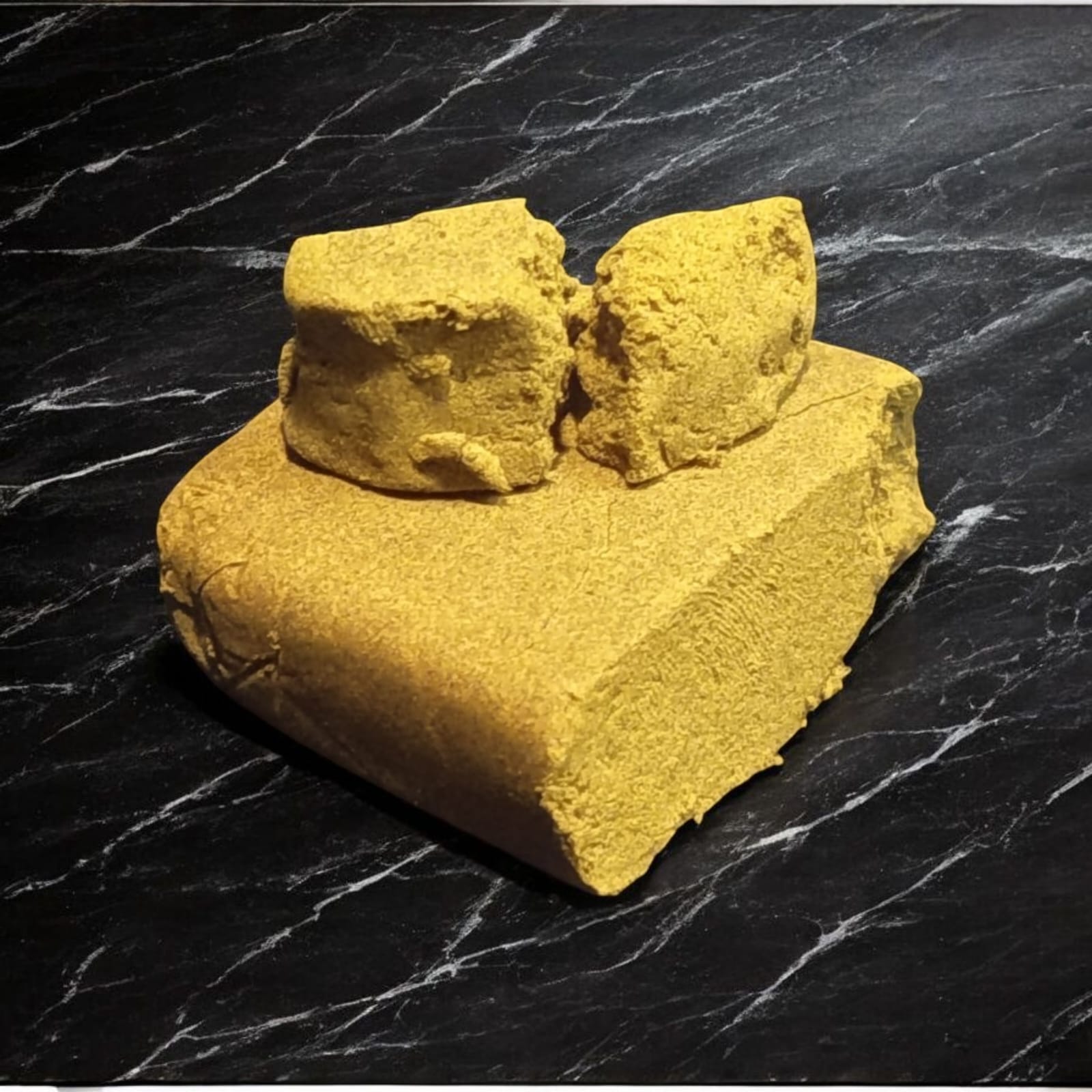 Bloc et morceaux de résine de CBD jaune, à texture friable, posés sur une surface noire marbrée.
